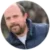 A man with a beard and short hair, identified as Alex Bye, wearing a blue quilted jacket and a white shirt, is outdoors. The image is in a circular frame.
