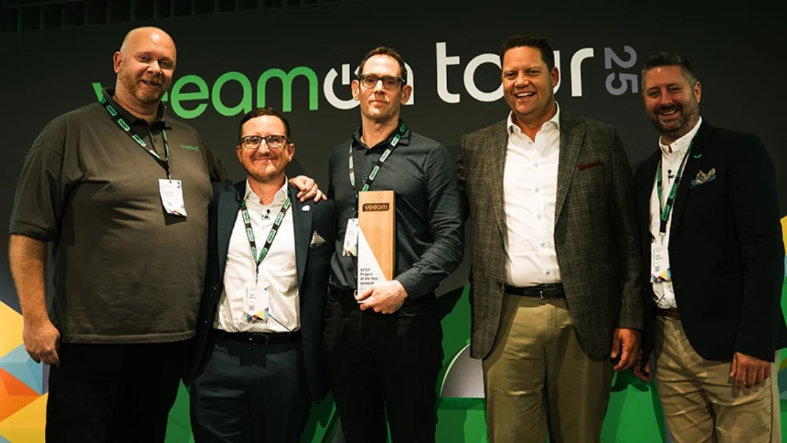 Dominic McLoughlin and Ant Bucknor on-stage with Veeam Executives smiling at a Veeam event, with Dominic in the centre holding a wooden award. They are wearing business attire and event badges, standing in front of a backdrop that reads Veeam on Tour 25.
