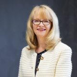 A smiling woman, identified as Nicola Downing, with blonde hair and glasses wears a cream textured jacket with gold buttons over a black blouse, sitting in front of a dark grey background.