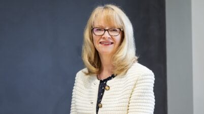 A smiling woman, identified as Nicola Downing, with blonde hair and glasses wears a cream textured jacket with gold buttons over a black blouse, sitting in front of a dark grey background.