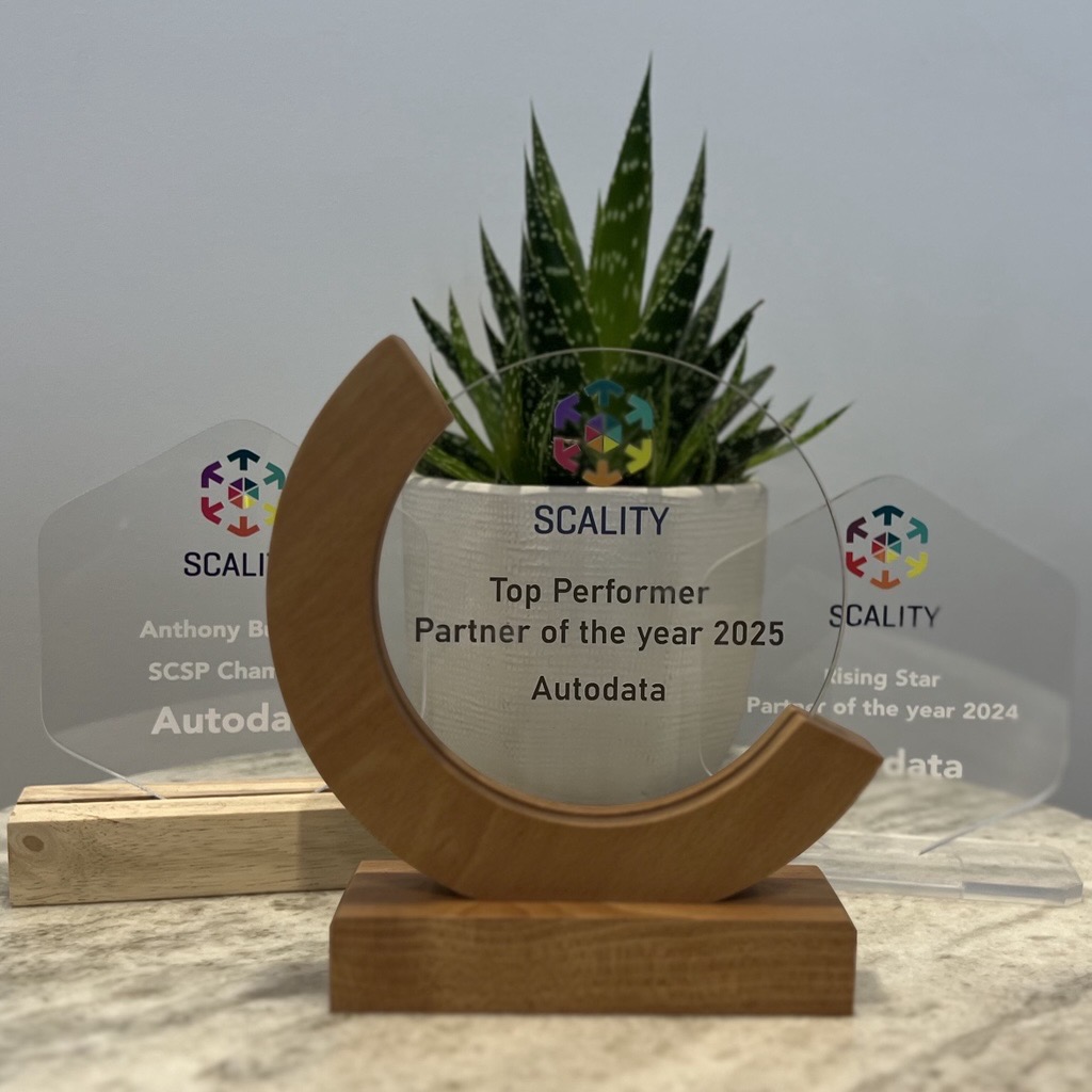 Three glass awards with colourful logos are displayed in front of a green potted succulent on a wooden surface. The central award reads Top Performer Partner of the year 2025 Autodata.