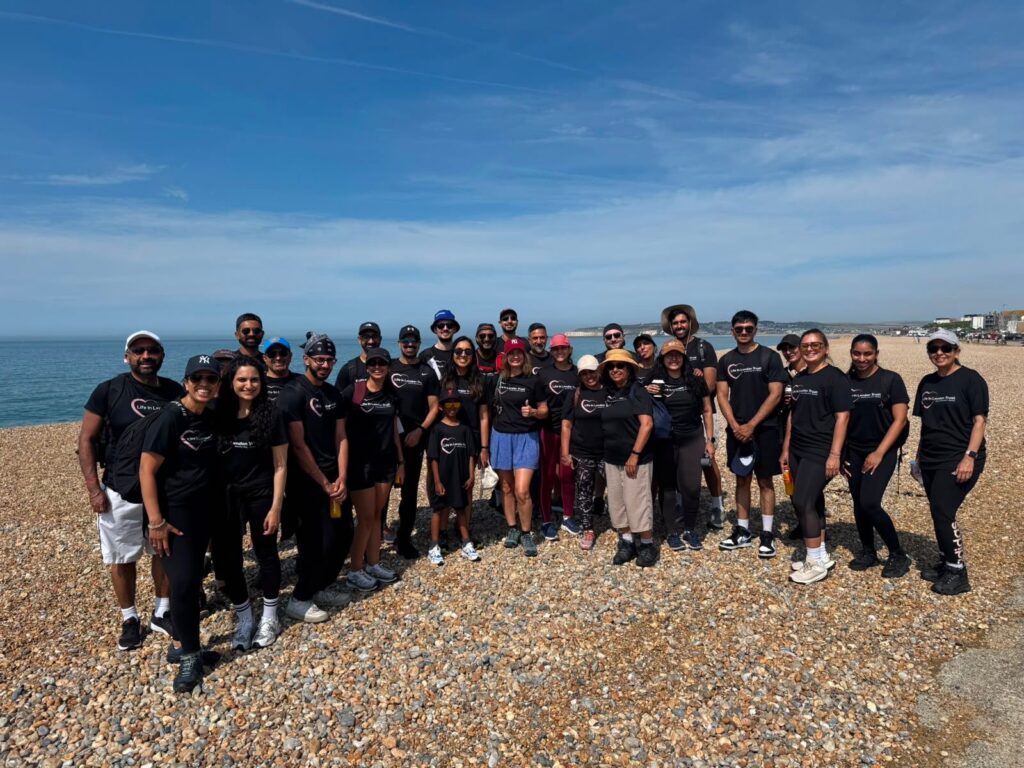 A group of people wearing matching black shirts stand and smile on a rocky beach under a blue sky, with the sea in the background, capturing a joyful moment reminiscent of life in London’s vibrant social scene.