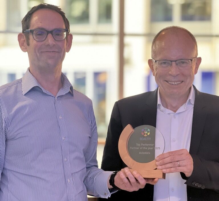 Two men stand indoors, smiling at the camera. One man holds a wooden and glass award plaque that reads SCALITY Partner Programme Awards 2023. The background shows large windows with a blurred cityscape.